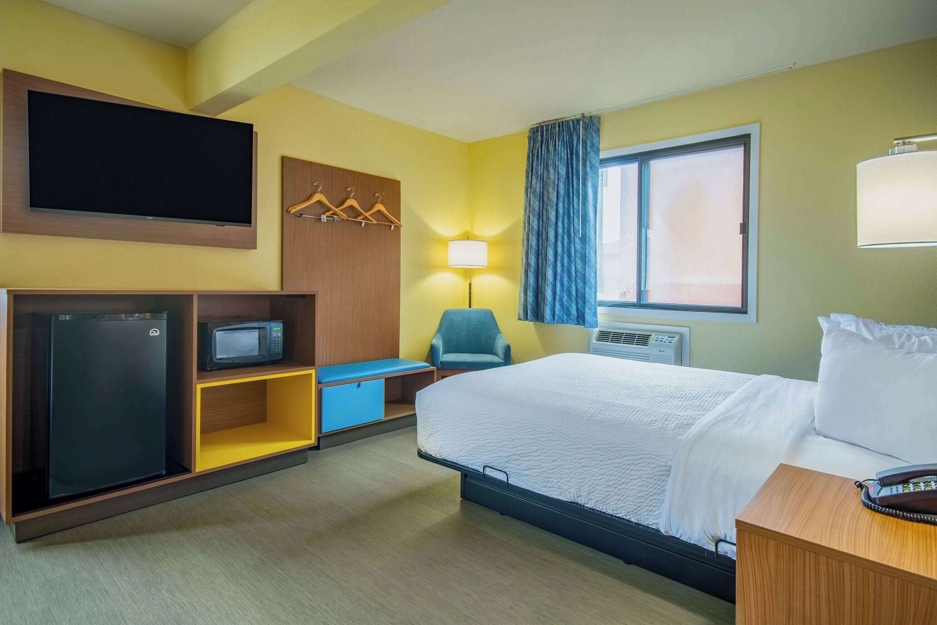 A hotel room with a bed, a TV mounted on the wall, a microwave, a small desk, a chair, and a window with curtains.