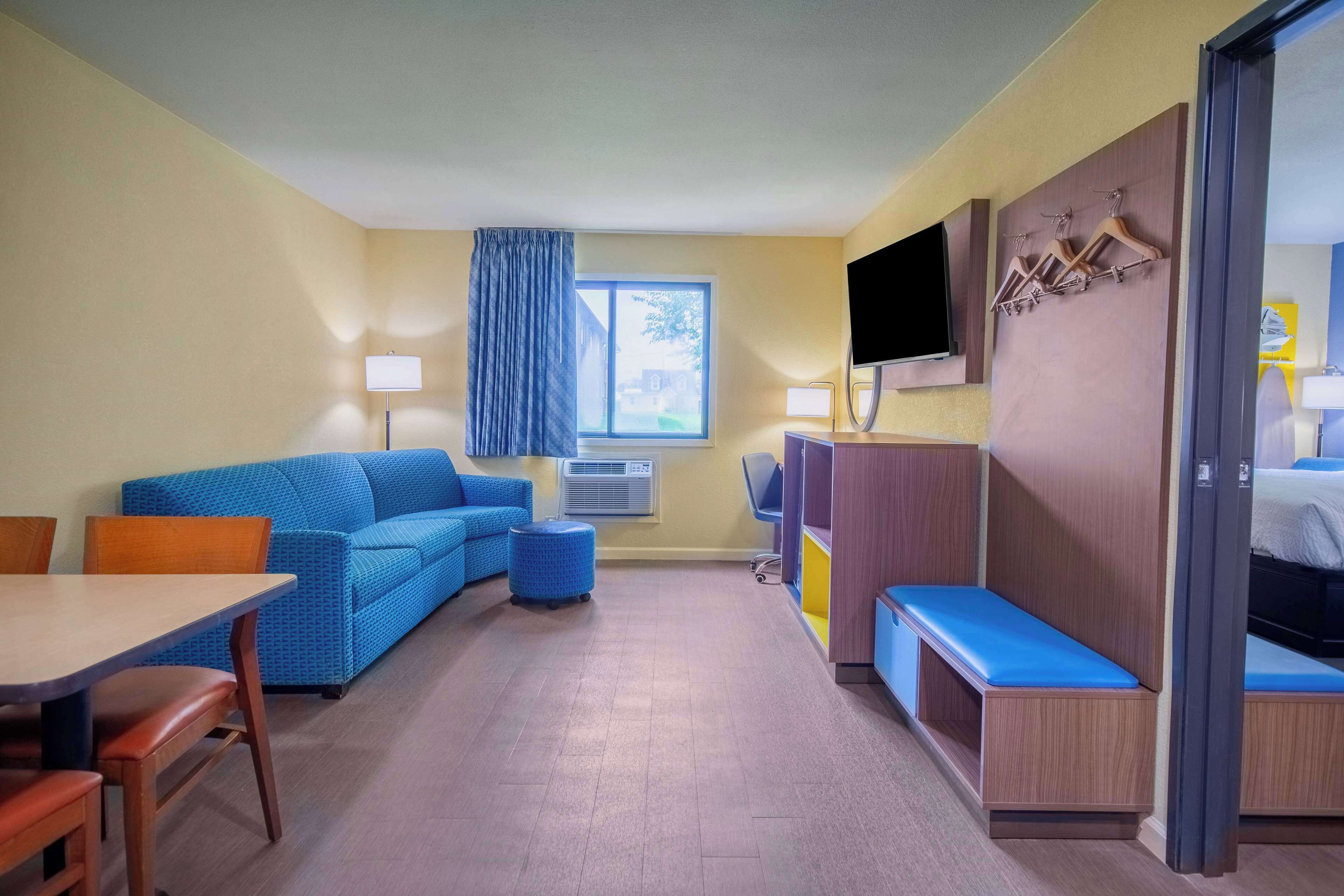 A hotel room with a blue sofa, a wooden table, a TV, a desk, a bench, and a wardrobe with a mirror.