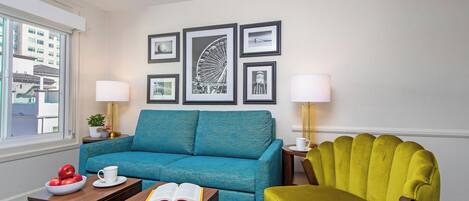 Une pièce avec un canapé bleu, un fauteuil vert, une table basse avec un livre dessus, et des tableaux encadrés accrochés au mur.
