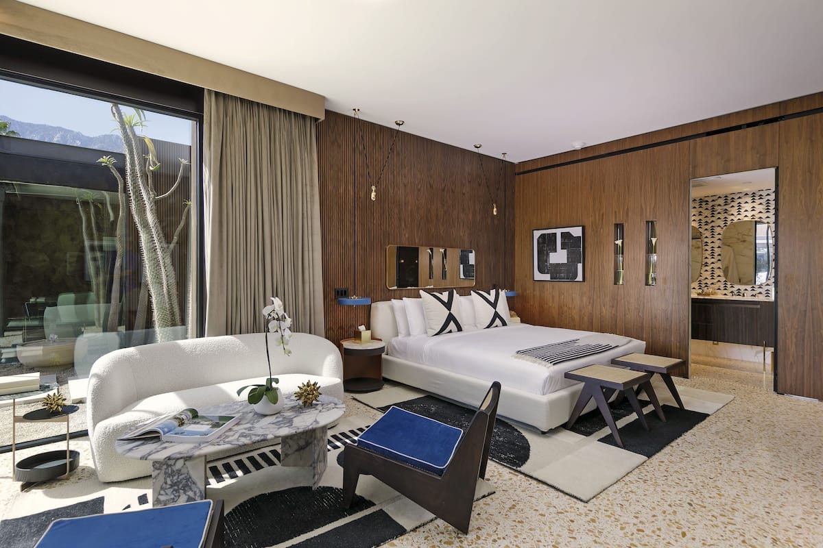 A modern hotel room with a large bed, a sofa, a small table, and a chair. There is a window with a view of mountains and a plant on the windowsill.