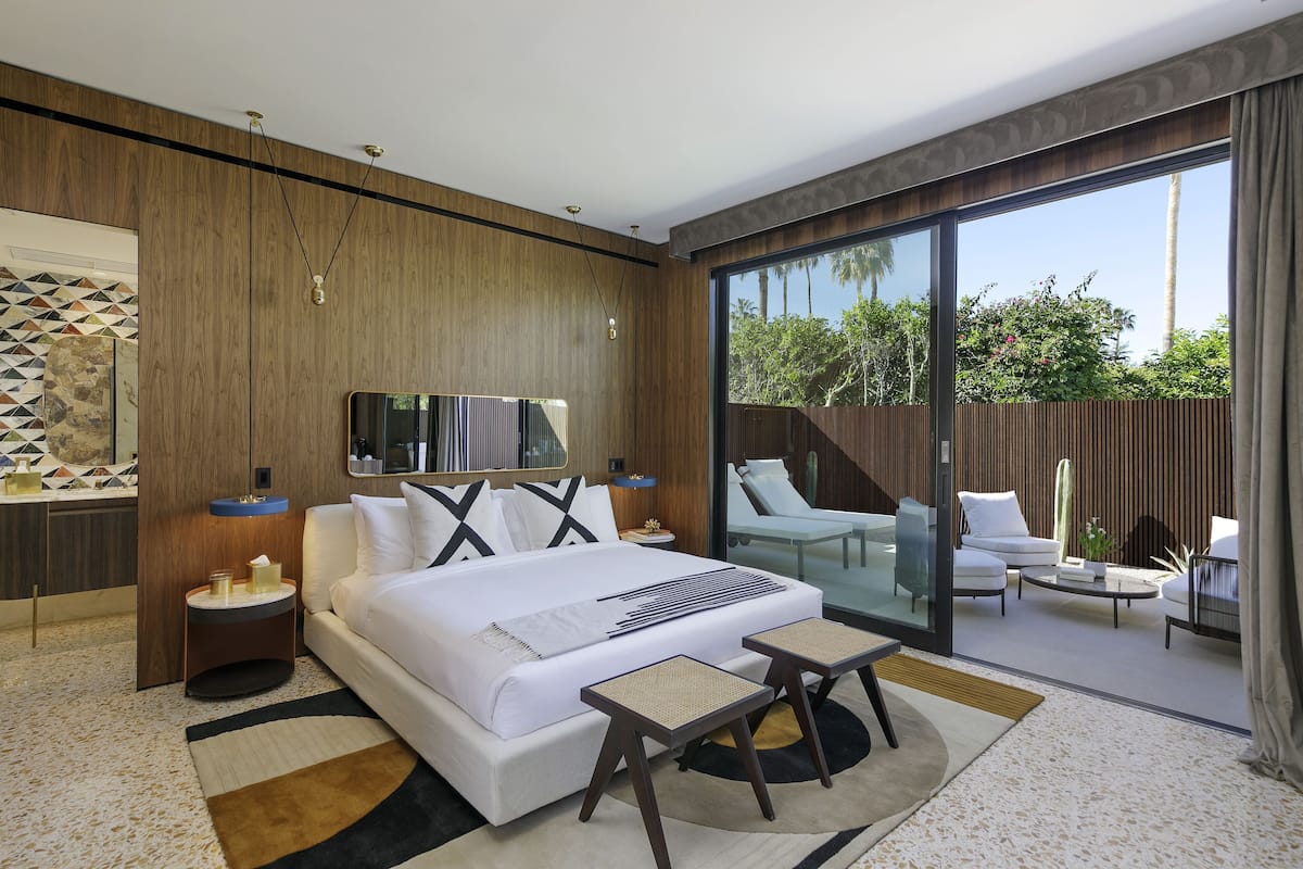 A modern hotel room with a large bed, a glass door leading to a balcony with seating, and a large window offering a view of greenery.