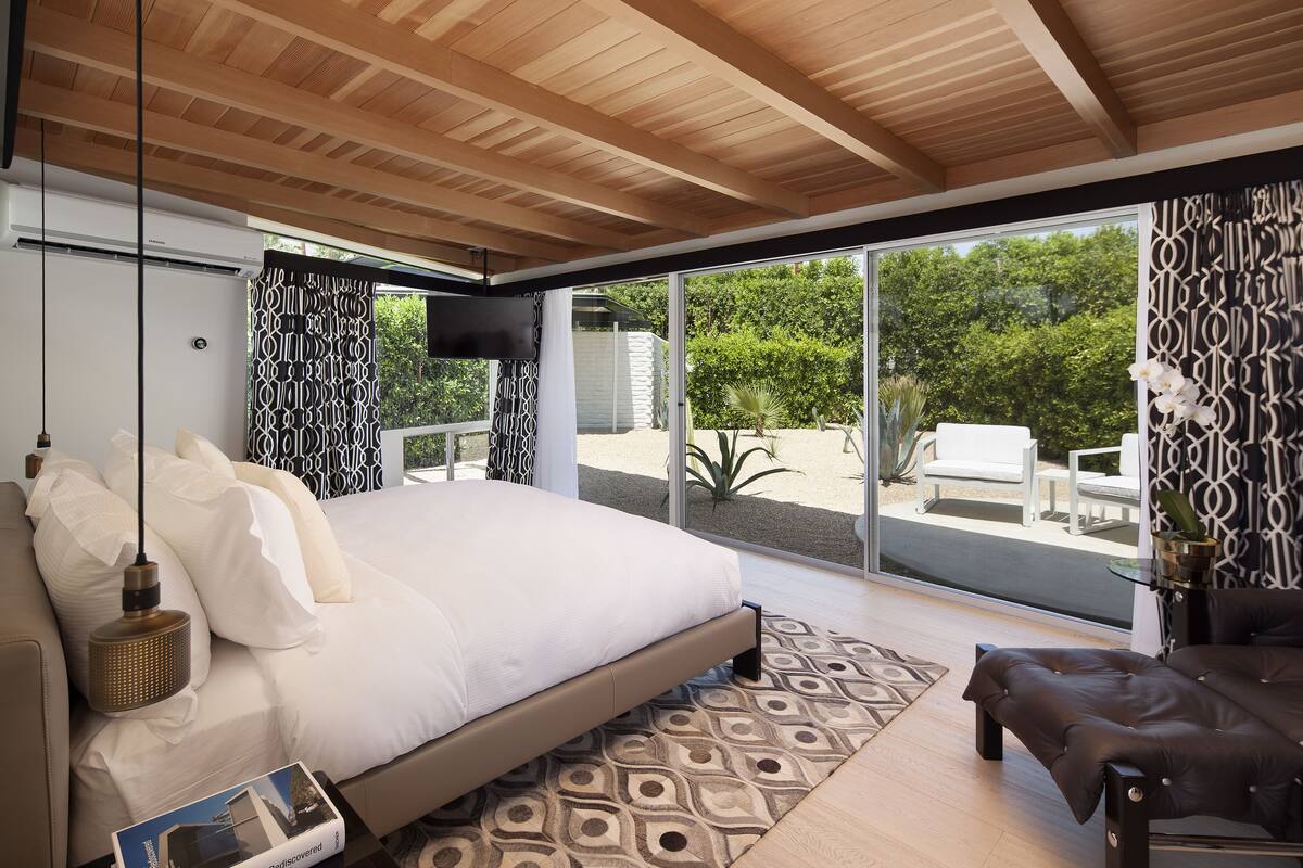 A modern bedroom with a large bed, a sofa, and a TV. It features a wooden ceiling, a large window, and a patterned rug.