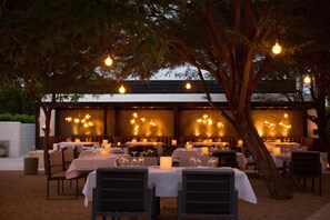 An outdoor dining area with tables set under a tree, surrounded by a wooden pergola with hanging lights.