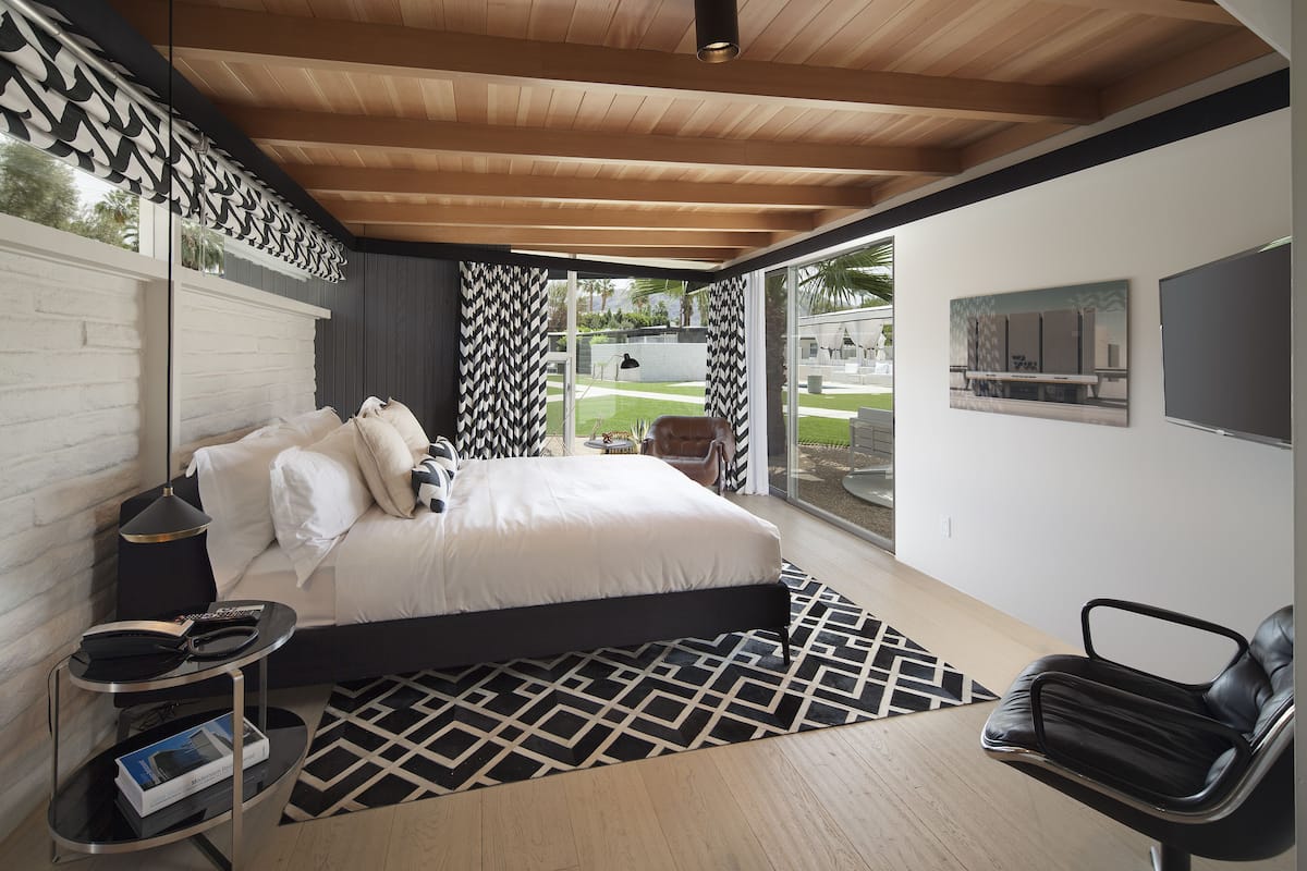 A modern bedroom with a large bed, a black and white patterned rug, a black chair, and a small side table with a book and remote control.