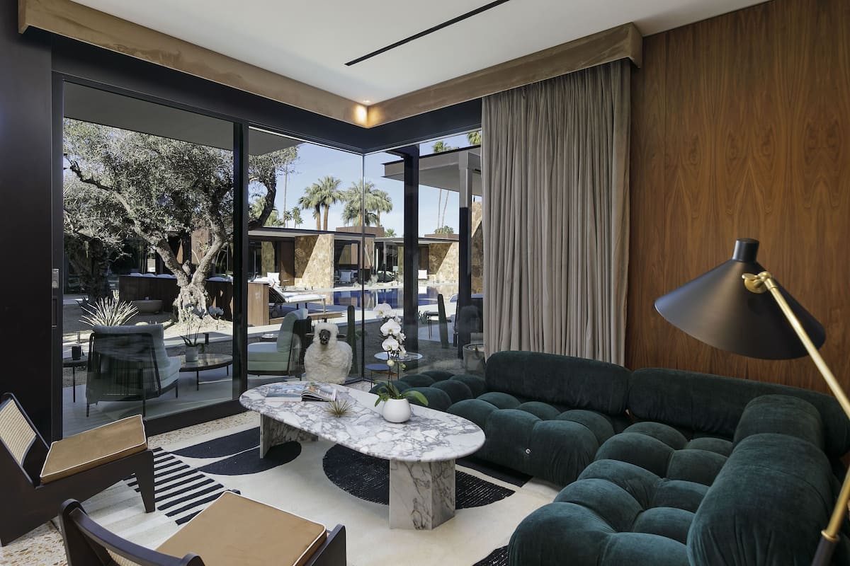A modern living room with a glass wall, a plush green sofa, a marble coffee table, and a large floor lamp.