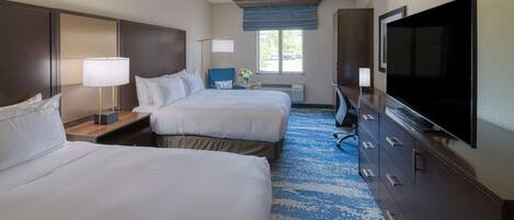 A hotel room with two beds, a desk with a lamp, a dresser, a television, and a large window with a view of the outside.