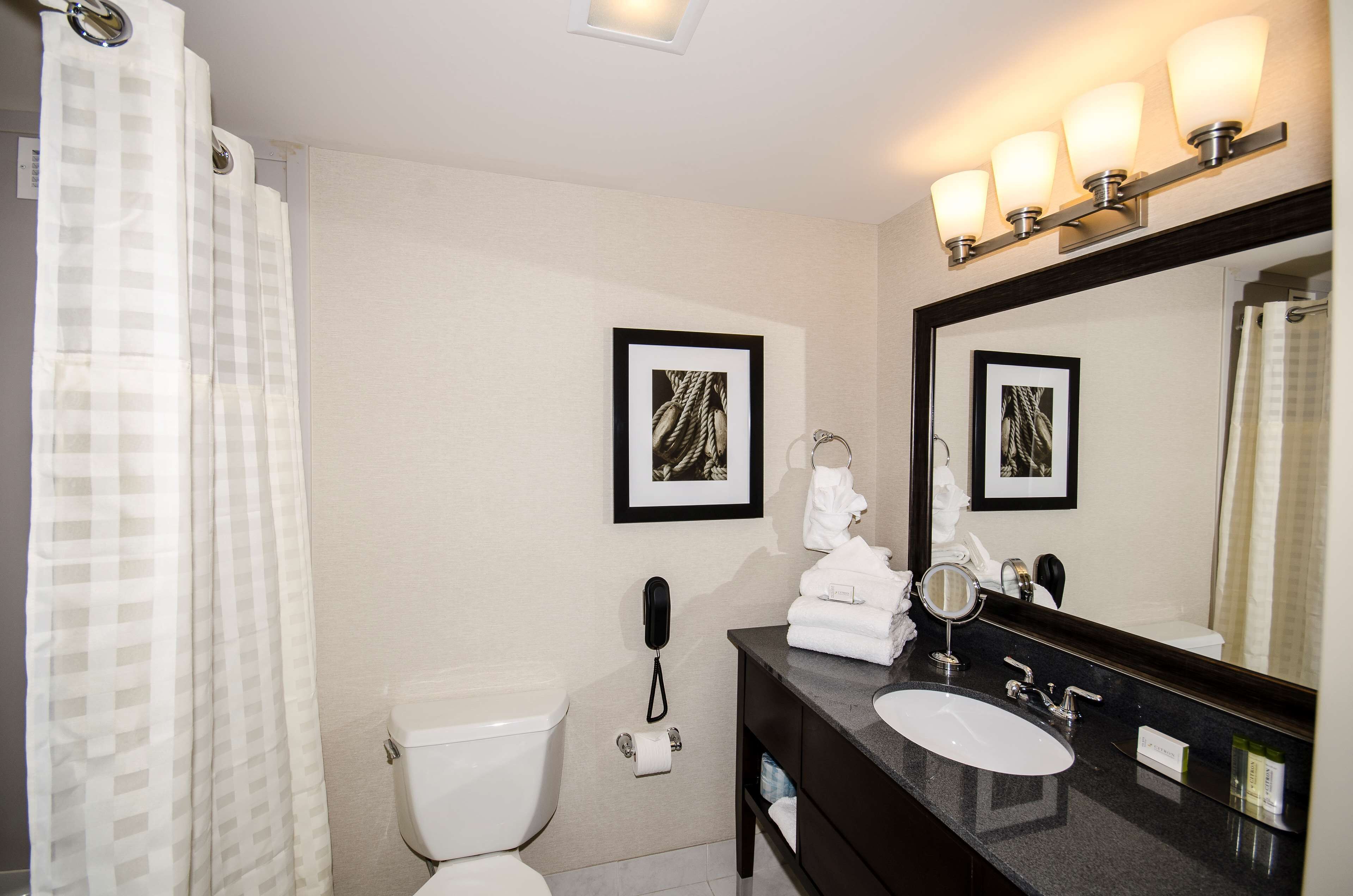 A bathroom with a toilet, sink, and mirror. There are framed pictures on the wall, a telephone, and a shower curtain.