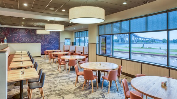 A modern dining area with round tables and chairs, large windows offering a view of a bridge and river, and a patterned wall. - DoubleTree by Hilton Hotel Port Huron (Port Huron)