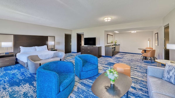A hotel room with a bed, two blue armchairs, a coffee table, a desk, and a kitchenette.