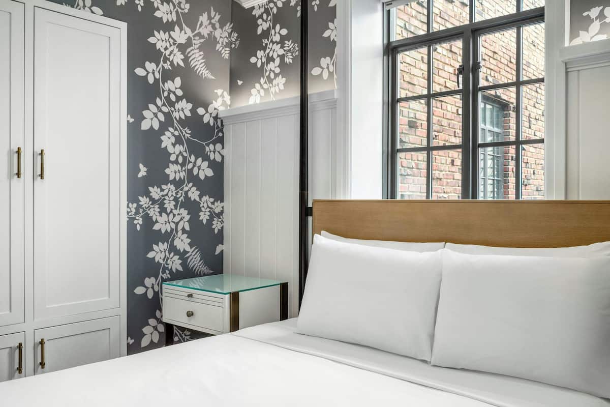 Une chambre avec un grand lit, une literie blanche, une tête de lit en bois, une table de chevet avec un plateau en verre et un mur orné d’un motif floral.