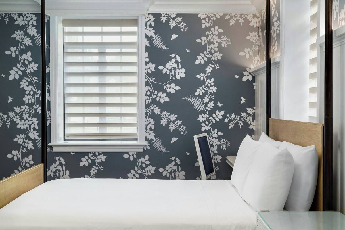 Une chambre à coucher avec un lit, une fenêtre avec des stores et un papier peint à motifs floraux.