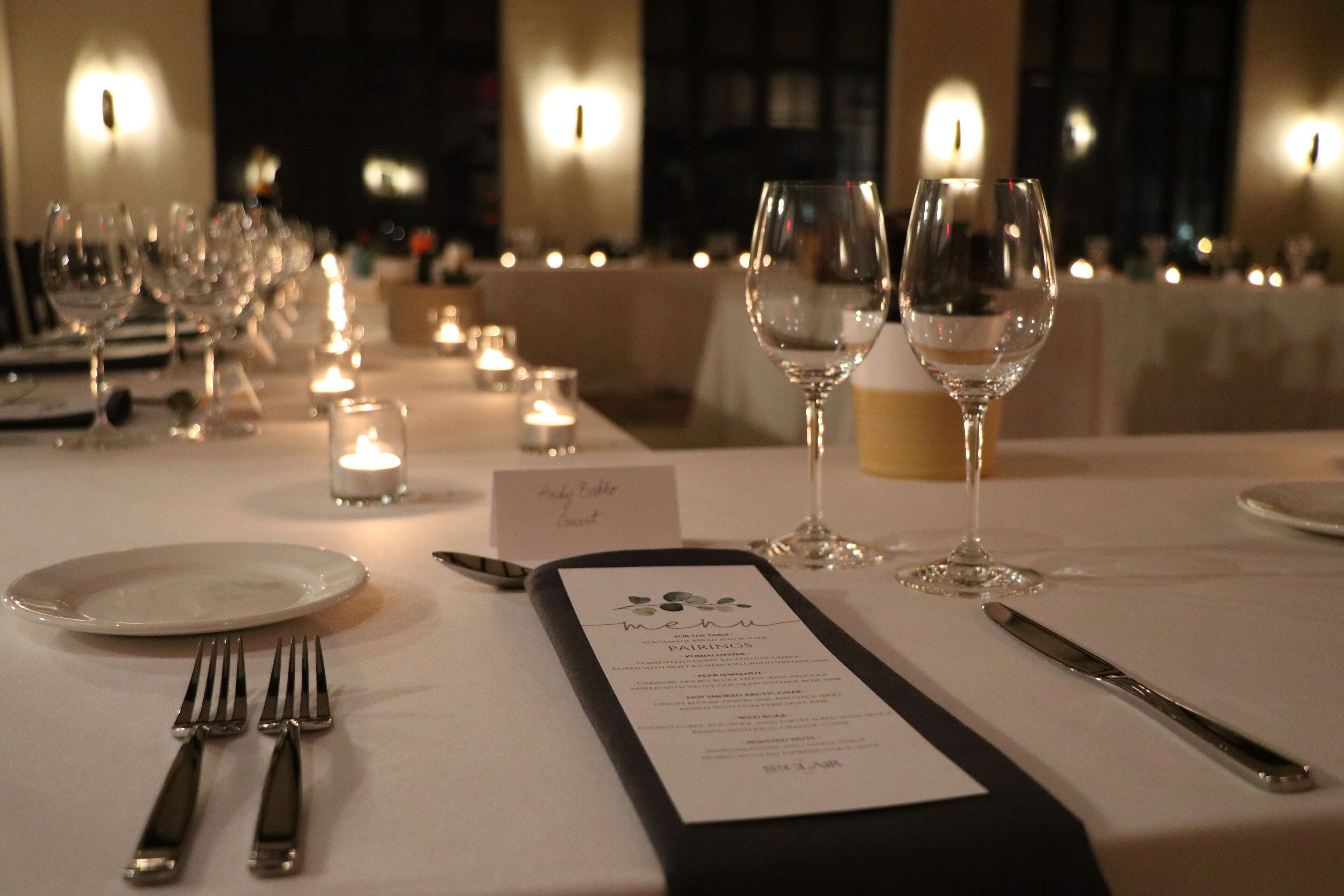 Una mesa de comedor puesta con platos, cubiertos, copas de vino y una carta.