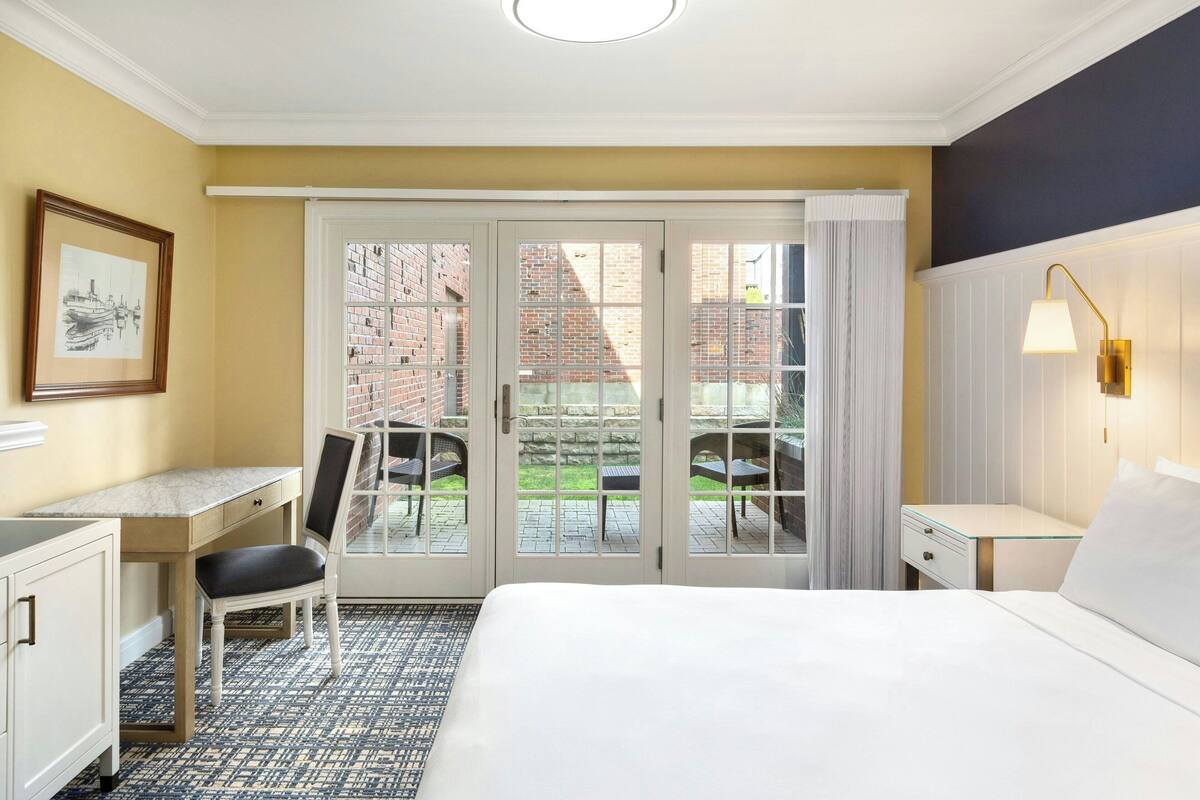 Une chambre d’hôtel avec un lit, un bureau, une chaise et une vue sur un immeuble en briques, visible à travers des portes vitrées.