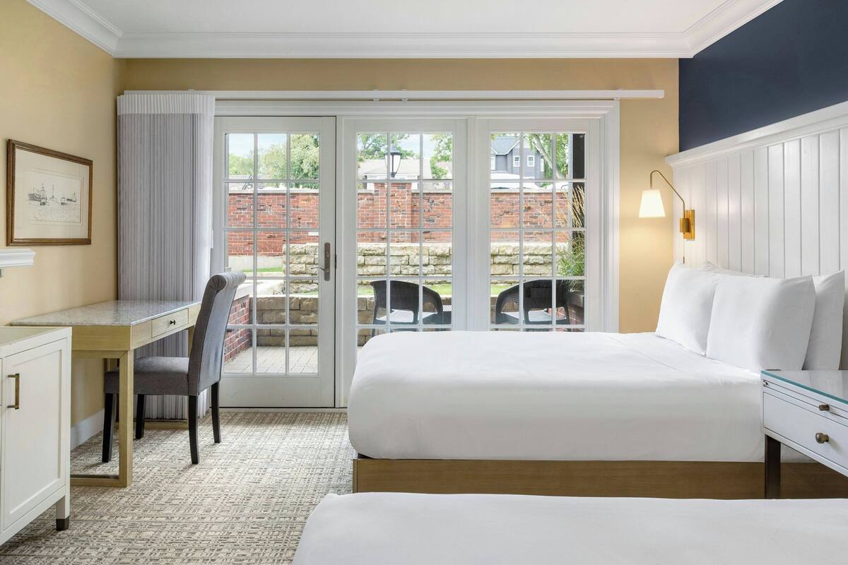 Une chambre d’hôtel avec un lit, un bureau et une chaise. Une grande fenêtre offre une vue sur un mur de briques et sur la verdure à l’extérieur.
