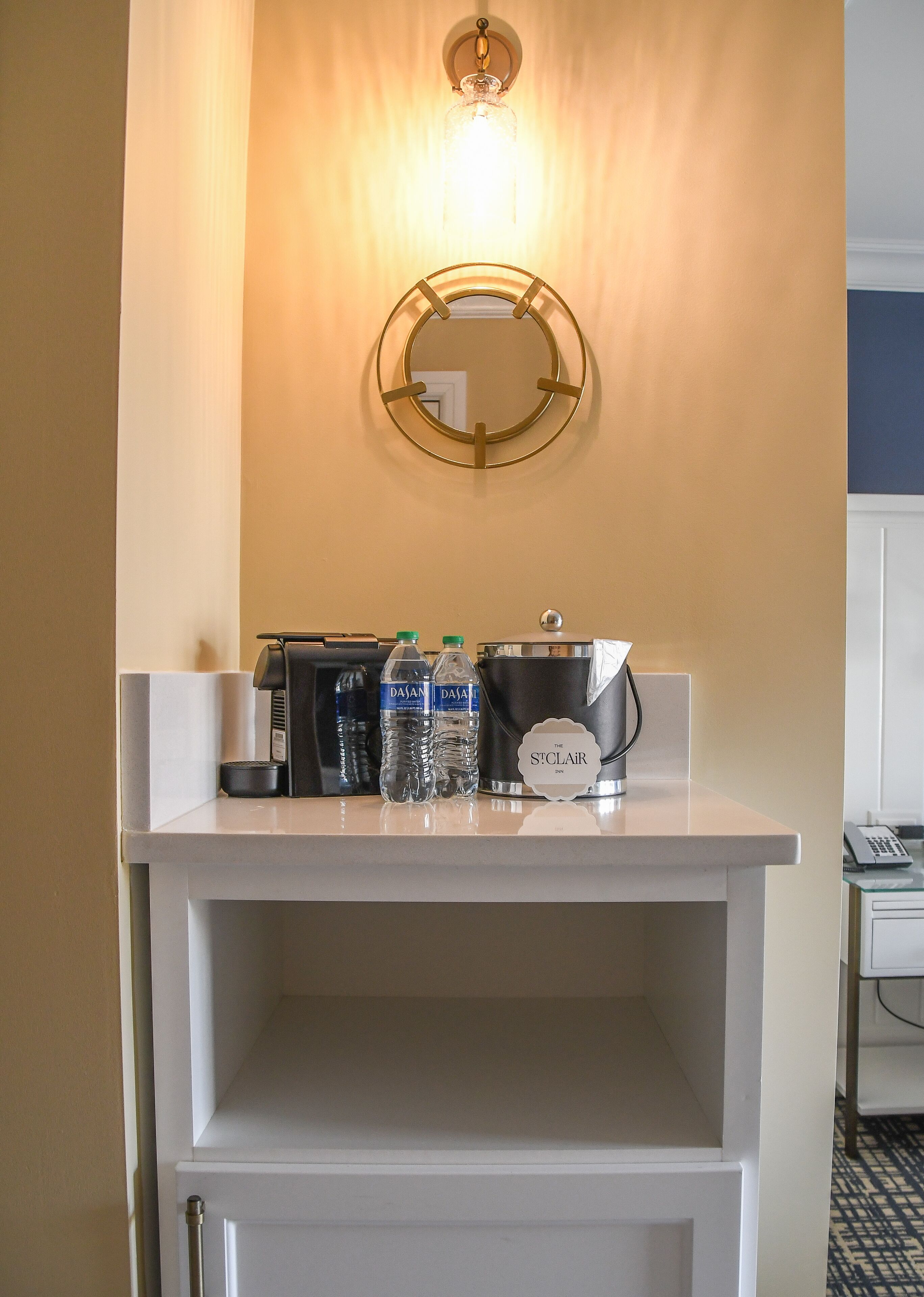 Een hotel receptie met een wit aanrecht, een koffieapparaat, twee waterflessen, een koffiekan en een spiegel met gouden lijst.