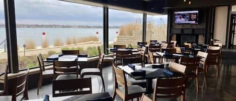 A restaurant with tables and chairs arranged in a spacious layout, large windows offering a view of the beach, and a flat-screen TV mounted on the wall.