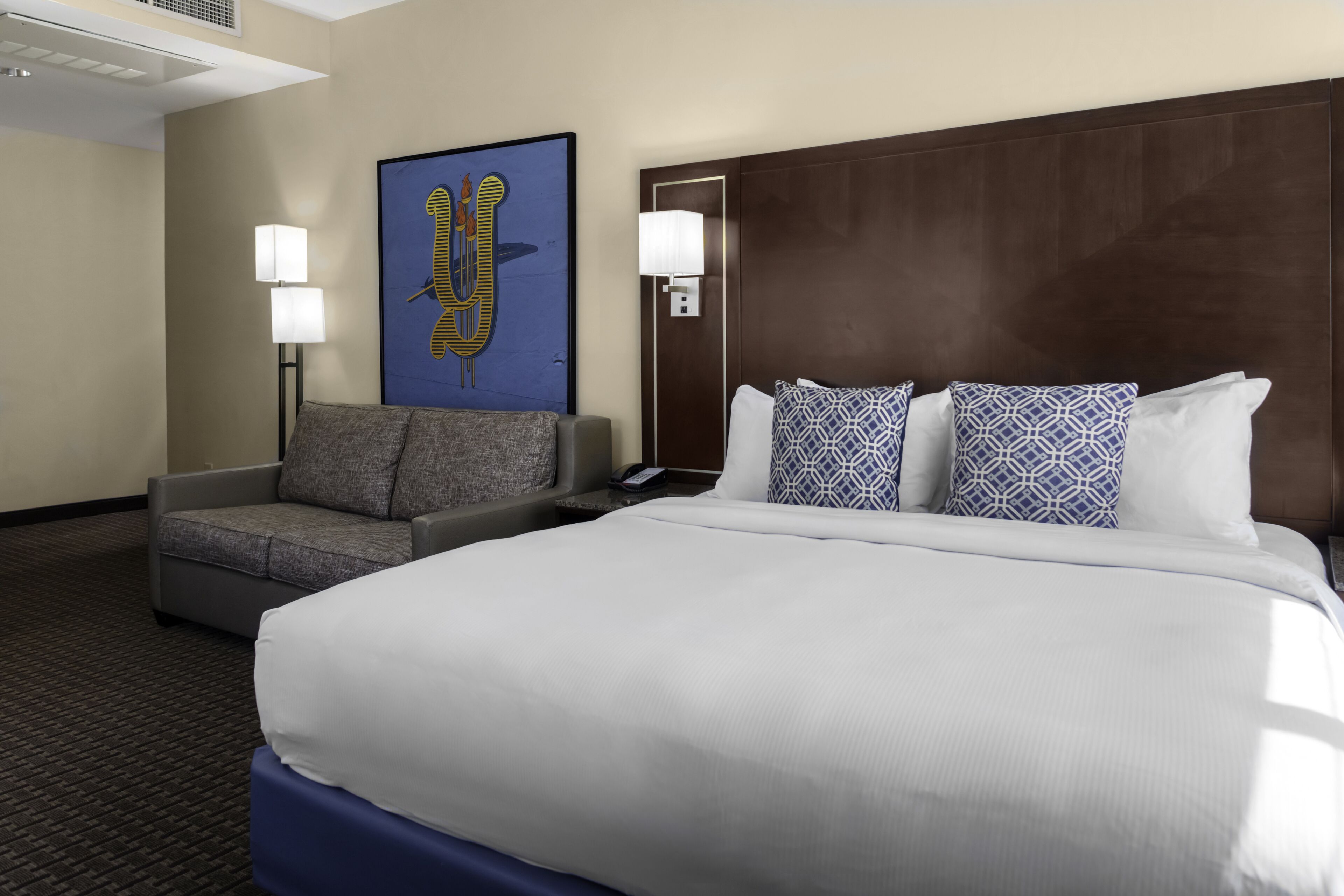 Quarto de hotel com uma cama grande, um sofá cinzento, duas luminárias fixadas na parede e uma obra de arte emoldurada.