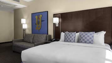Quarto de hotel com uma cama grande, um sofá cinzento, duas luminárias fixadas na parede e uma obra de arte emoldurada.
