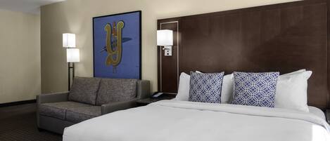 A hotel room with a large bed, a grey sofa, two wall-mounted lamps, and a framed artwork on the wall.