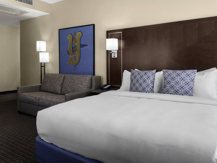 Quarto de hotel com uma cama grande, um sofá cinzento, duas luminárias fixadas na parede e uma obra de arte emoldurada.