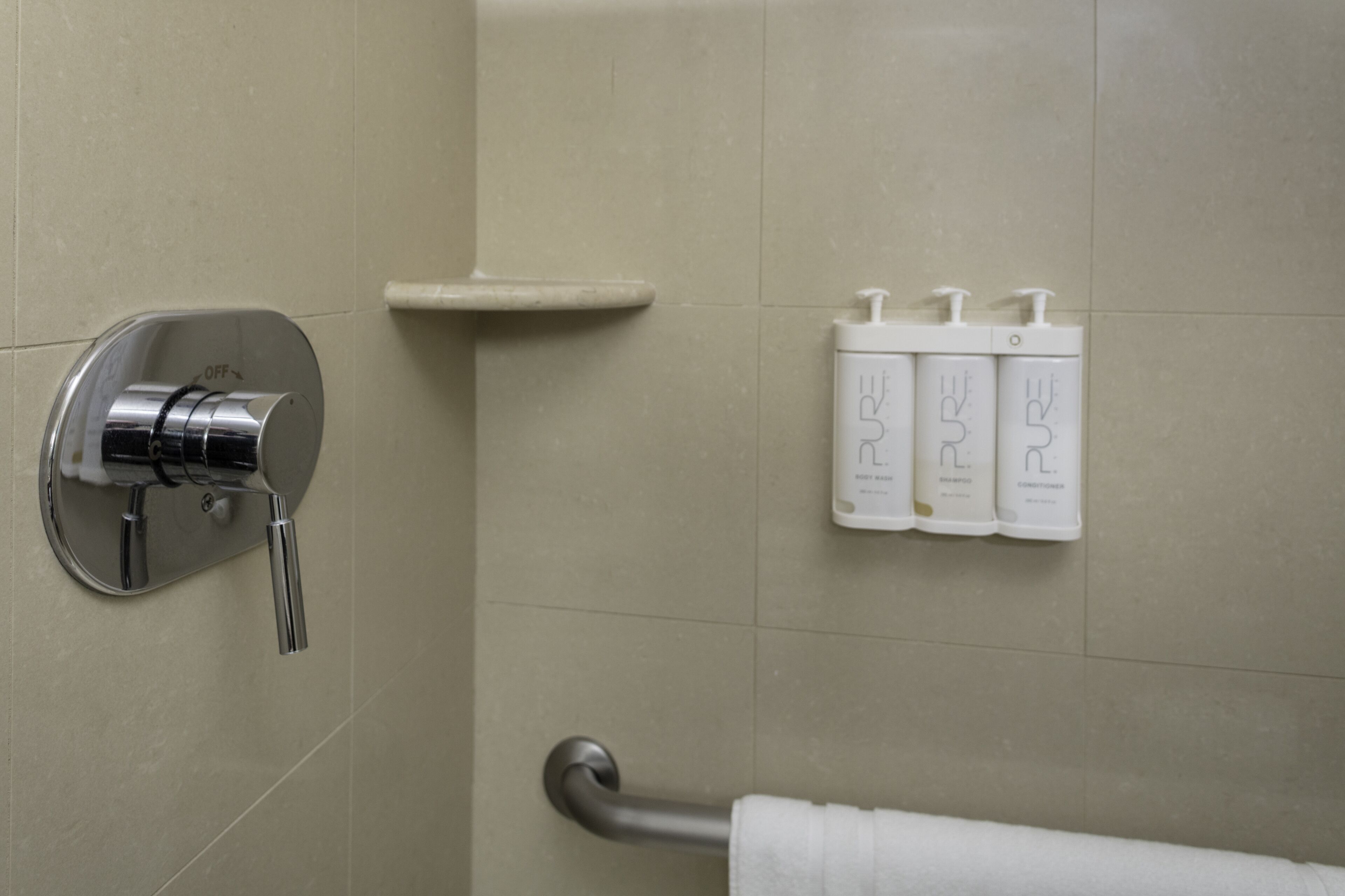 A bathroom with a shower control, a towel rack, and three bottles of Pure shampoo and conditioner.