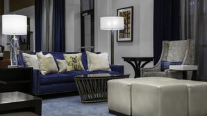 A modern living room with a blue sofa, white ottoman, and a grey armchair. There are two lamps on side tables, and a framed artwork on the wall. - New Haven Hotel (New Haven)