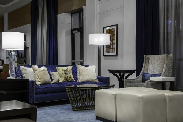 Una sala moderna con un sofá azul, un reposapiés blanco y un sillón gris. Hay dos lámparas sobre mesitas de noche y una obra de arte enmarcada en la pared.