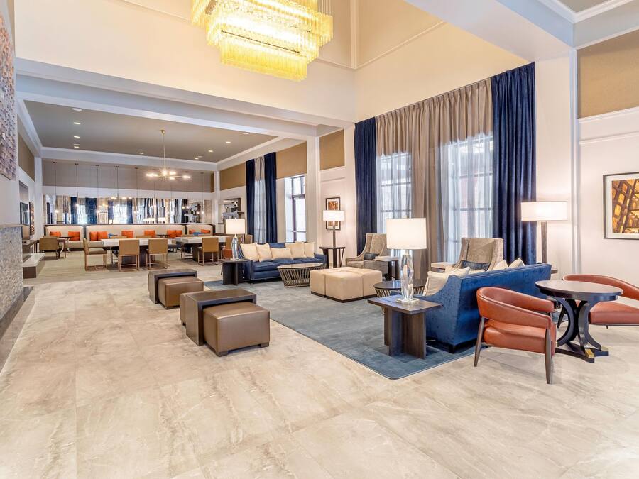 A spacious hotel lobby with a modern design, featuring a seating area with sofas and chairs, a dining area, and a large chandelier.
