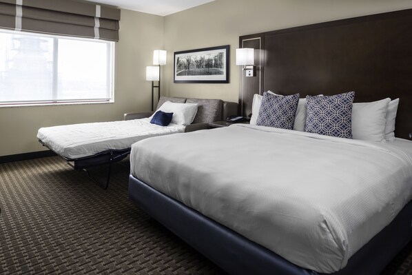 A hotel room with a large bed, two smaller beds, a headboard, a nightstand, a lamp, a framed picture on the wall, and a window with blinds.