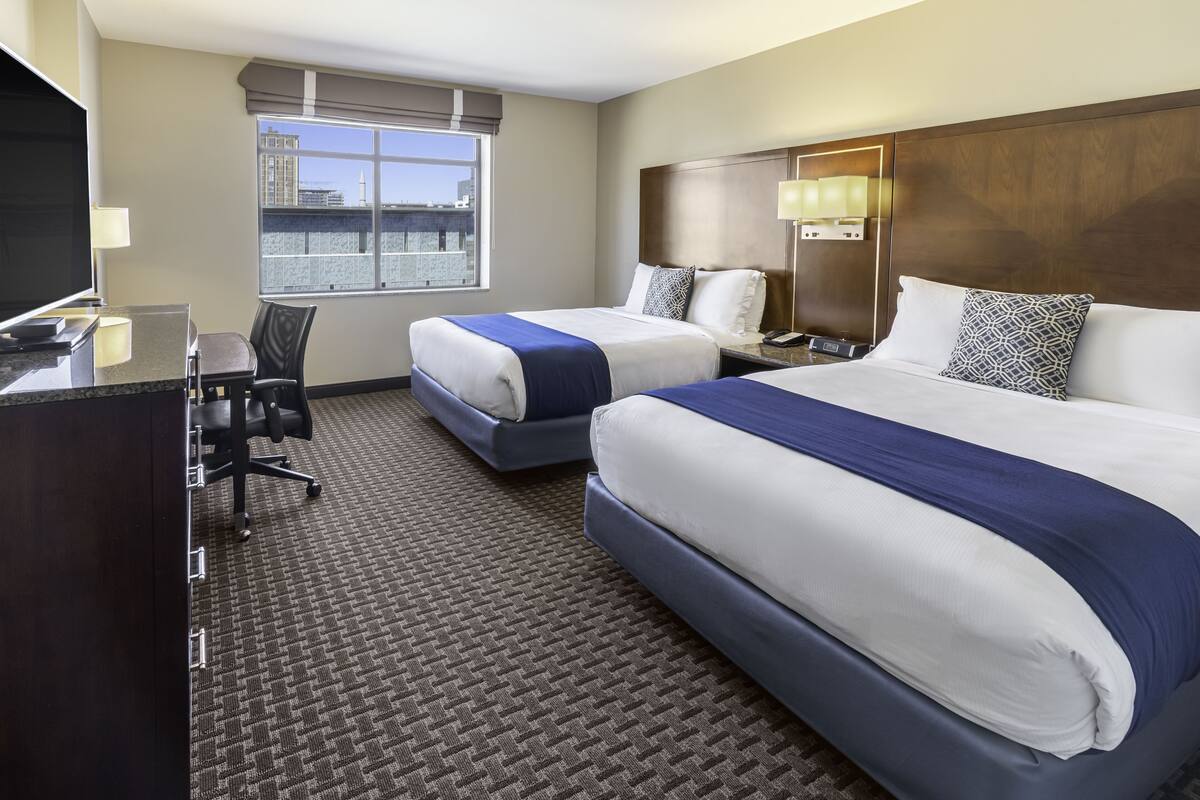 A hotel room with two beds, a desk, a chair, a TV, and a window with a city view.