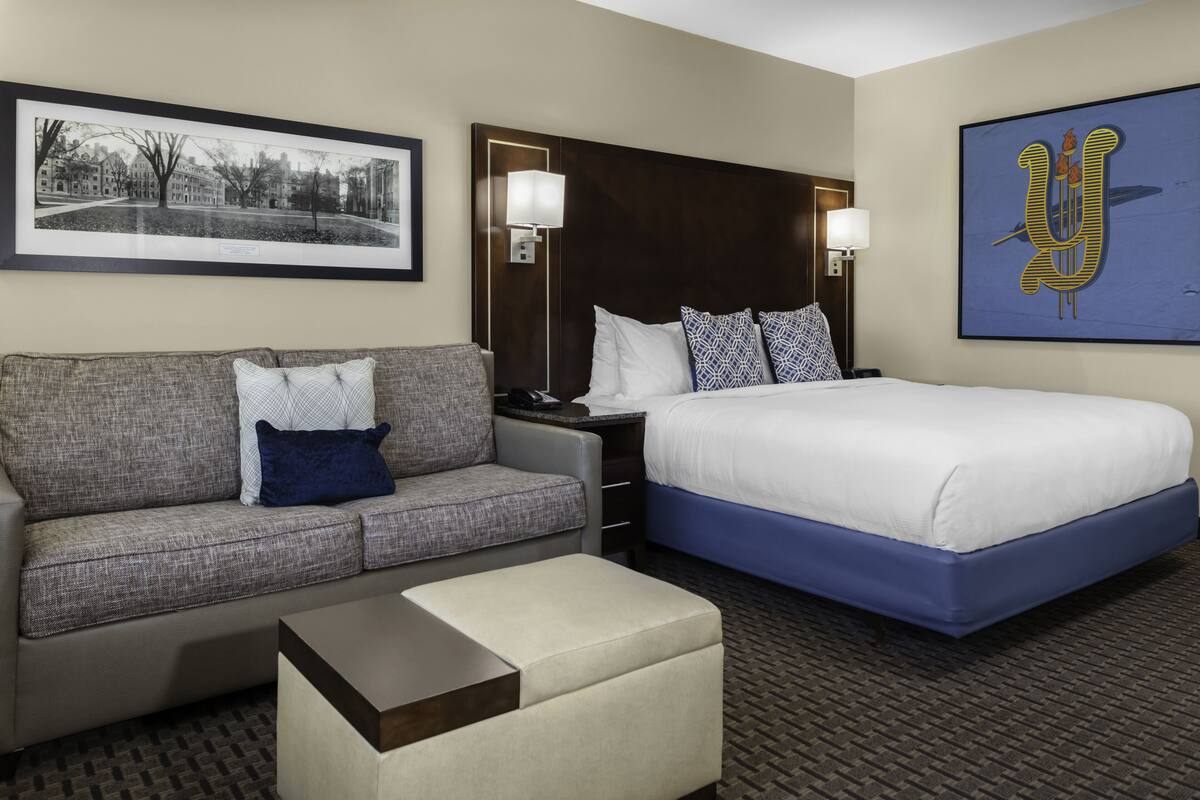 A hotel room with a bed, a sofa, a nightstand, a lamp, a framed picture on the wall, and a carpeted floor.
