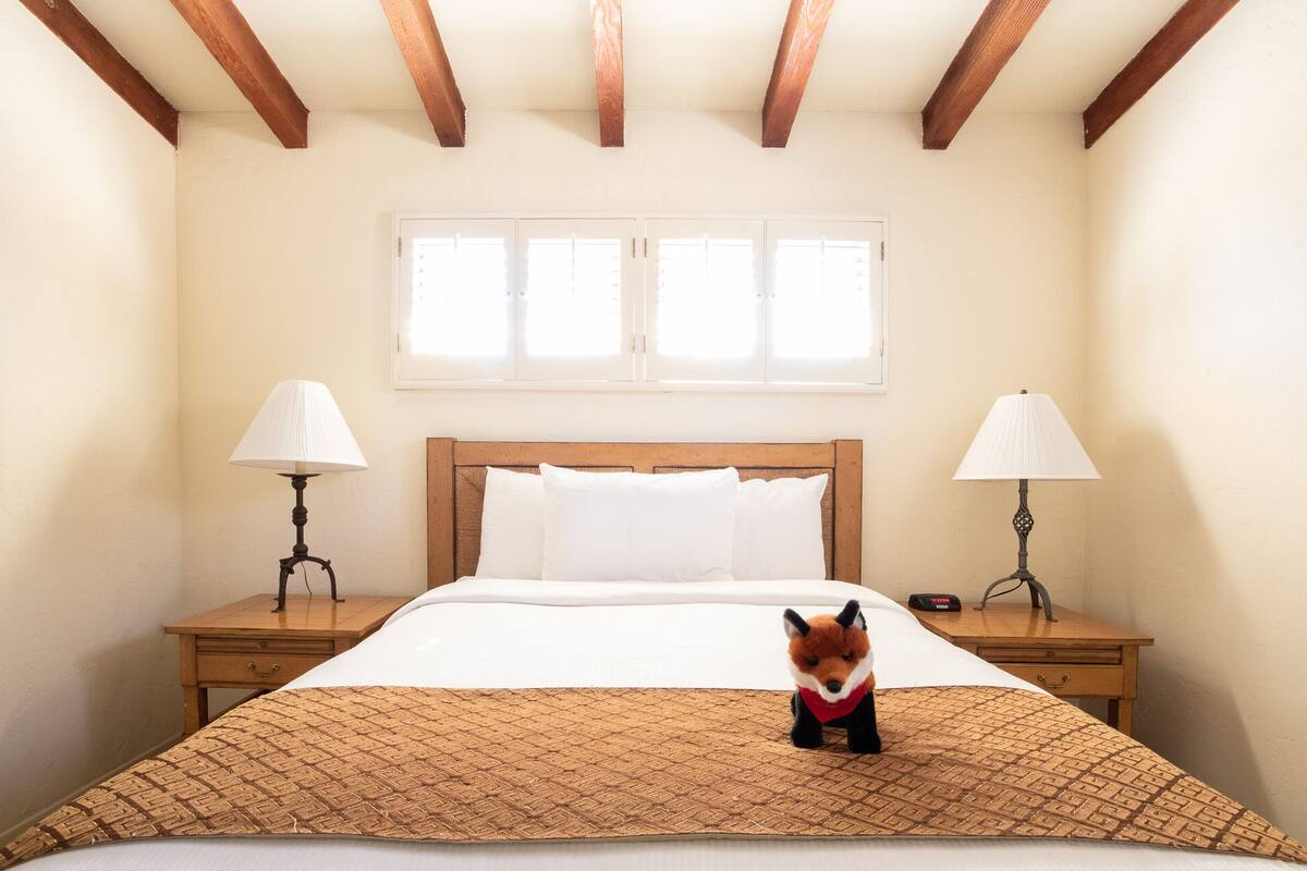 Ein Schlafzimmer mit einem Holz-Kopfteil, einem Bett mit weißer Bettwäsche, zwei Nachttischlampen, einem Fenster mit Jalousien und einem kleinen Fuchs-Plüschtier auf dem Bett.
