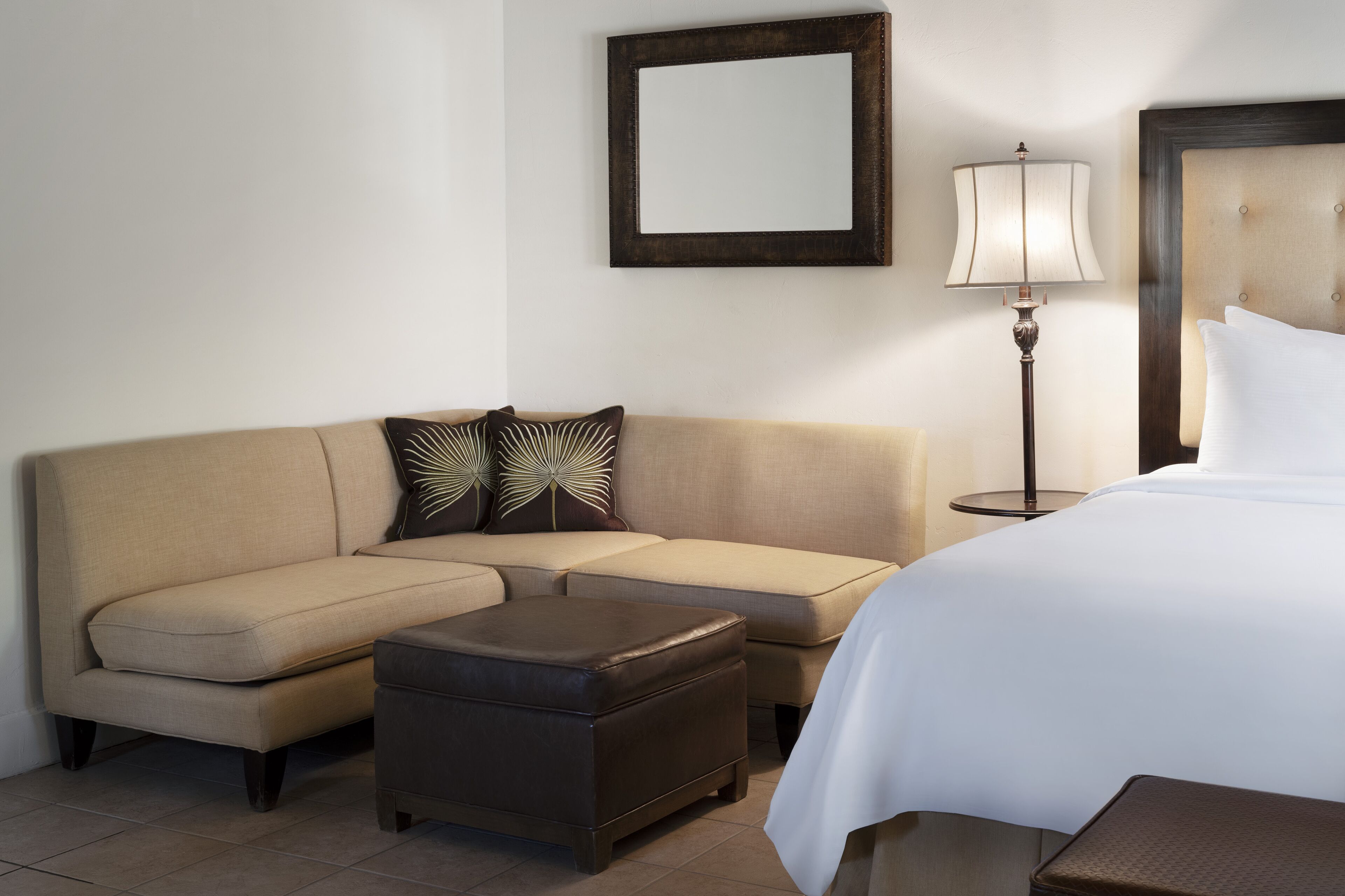 A hotel room with a beige sofa, a matching ottoman, a bed with white bedding, a bedside table, a lamp, and a framed mirror on the wall.