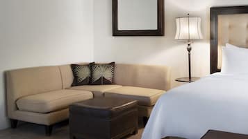 A hotel room with a beige sofa, a matching ottoman, a bed with white bedding, a bedside table, a lamp, and a framed mirror on the wall.