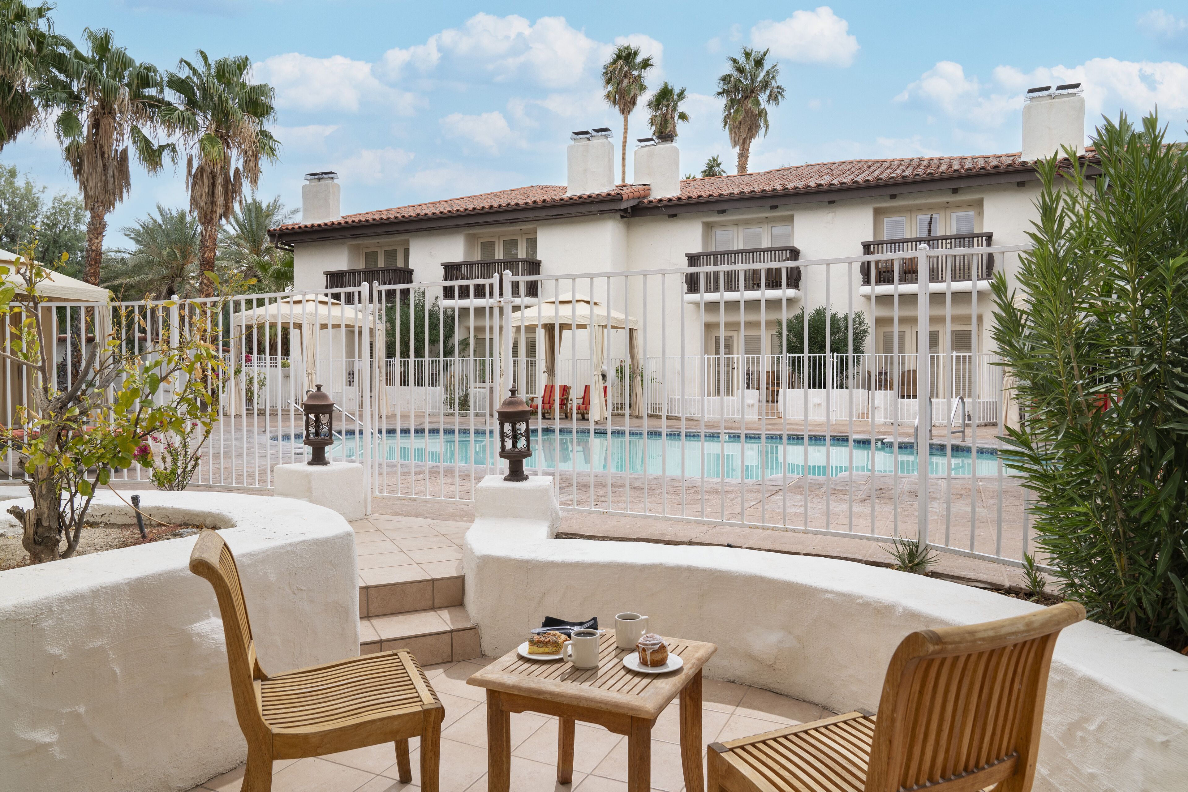 Zona de piscina con valla blanca, sillas de madera y una mesa con comida y bebidas.