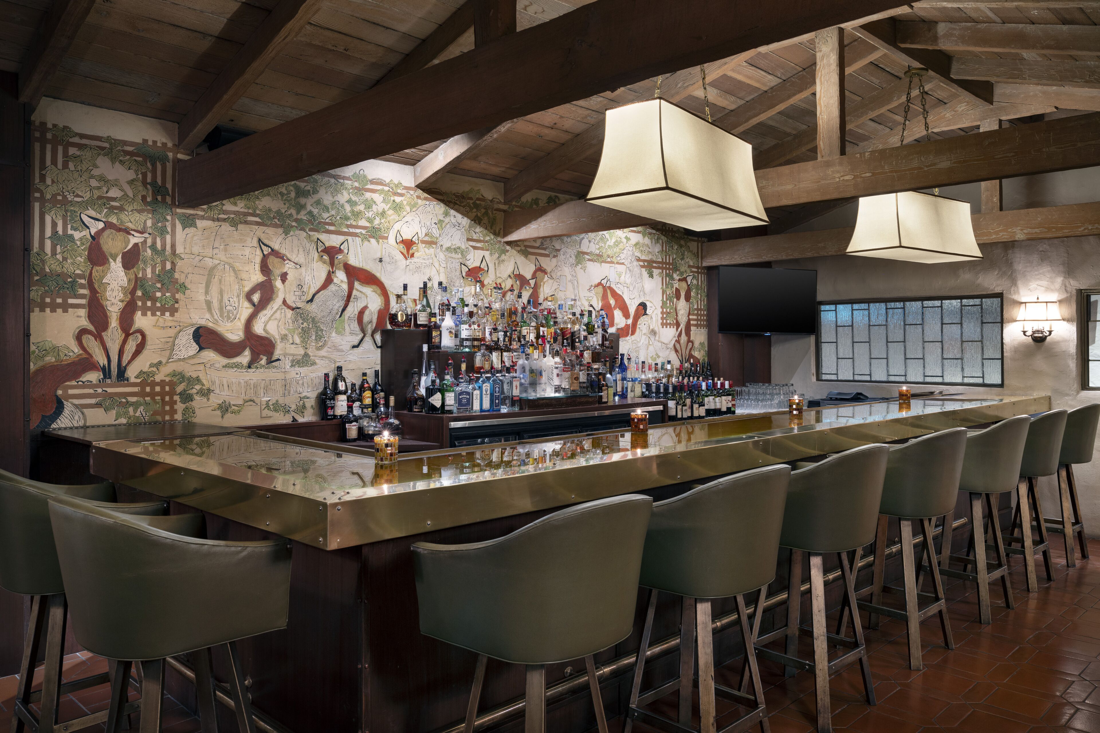 A bar area with a long counter, multiple stools, and a mural of foxes on the wall.