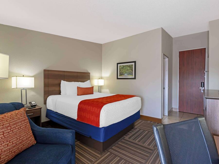 A hotel room with a bed, a desk, a chair, a lamp, and a door.