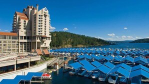 A marina with numerous boats docked under blue canopies, a large hotel building with multiple floors and balconies, and a forested hillside in the background. - The Coeur D'alene Golf and Spa Resort (Coeur d'Alene)