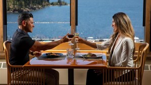 A dining table with two people toasting with wine glasses, overlooking a lake with a forested shoreline. - The Coeur D'alene Golf and Spa Resort (Coeur d'Alene)