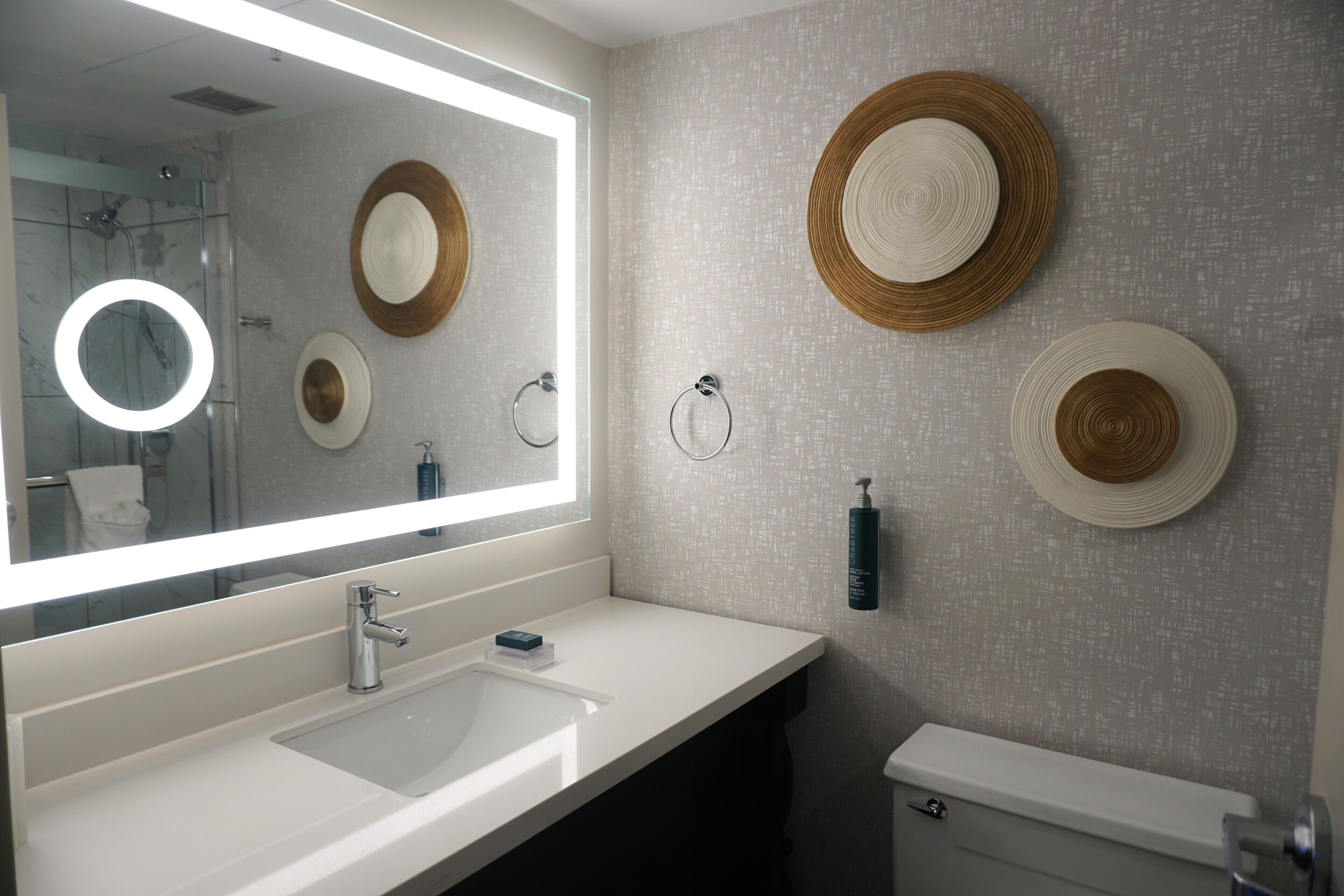 A modern bathroom with a large mirror, white sink, and toilet. There are decorative plates on the wall and a soap dispenser.