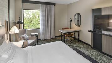 Ein Hotelzimmer mit Bett, Schreibtisch, Stuhl, kleinem Tisch, Lampe, Fernseher und einem Fenster mit Vorhängen.