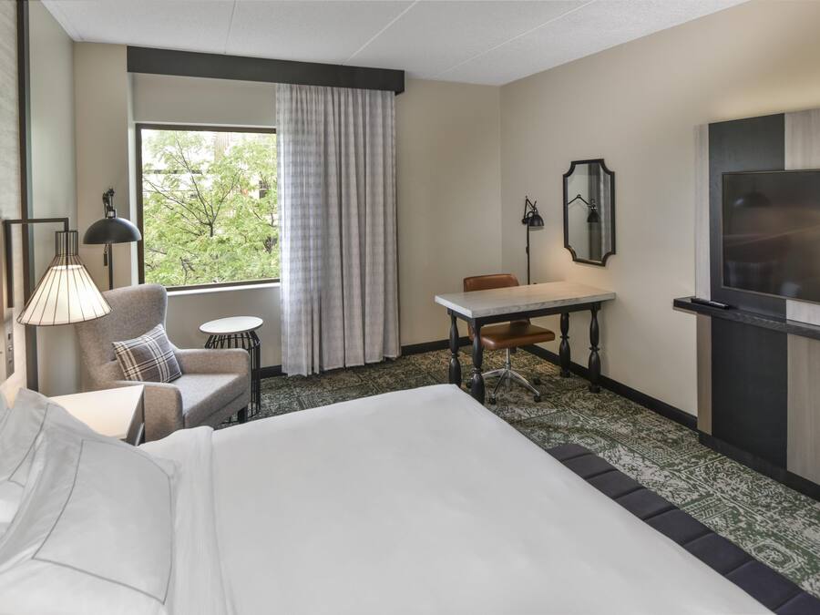 Ein Hotelzimmer mit Bett, Schreibtisch, Stuhl, kleinem Tisch, Lampe, Fernseher und einem Fenster mit Vorhängen.