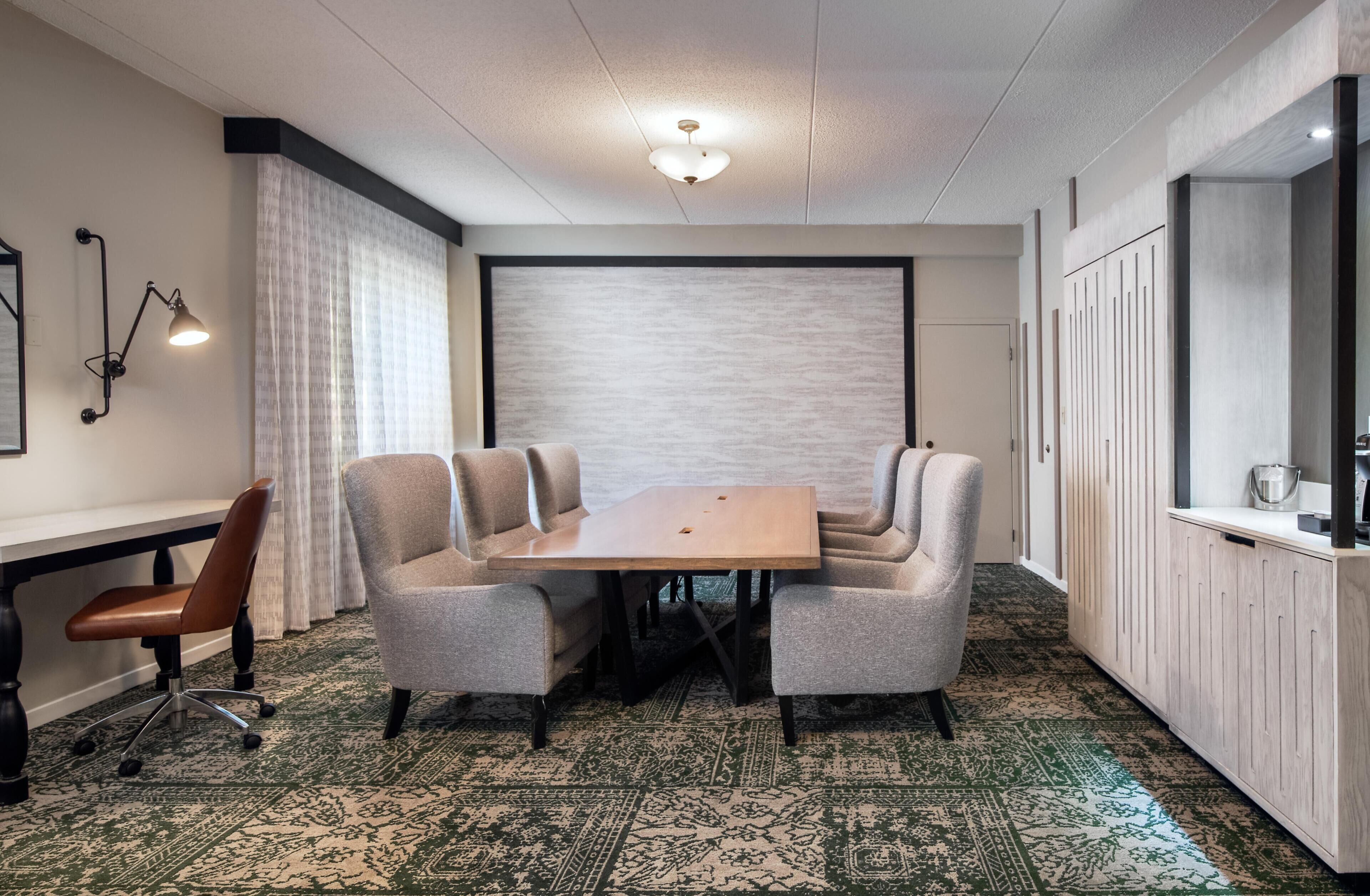 Un salón de conferencias con una mesa rectangular, cuatro sillas tapizadas, una silla de oficina individual, una alfombra estampada, un techo blanco con una lámpara, y una puerta blanca.