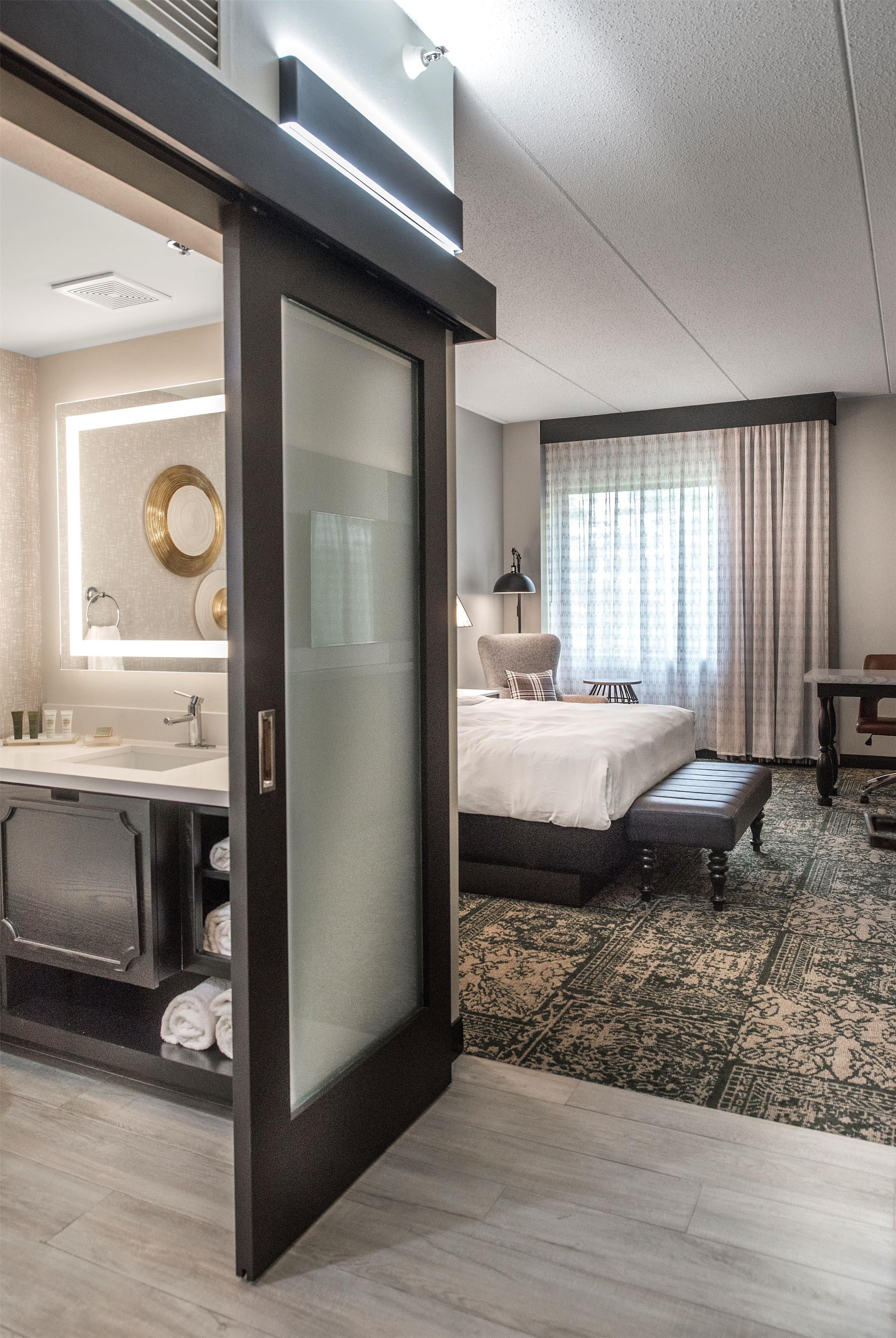 A hotel room with a large bed, a separate bathroom with a vanity, a desk, and a window with curtains.