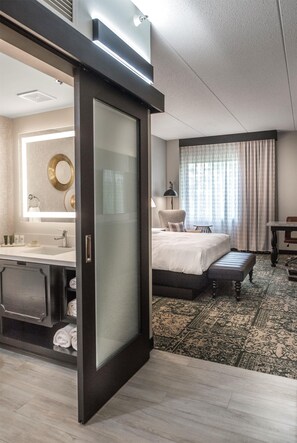 A hotel room with a large bed, a separate bathroom with a vanity, a desk, and a window with curtains. - Hilton Appleton Paper Valley (Appleton)