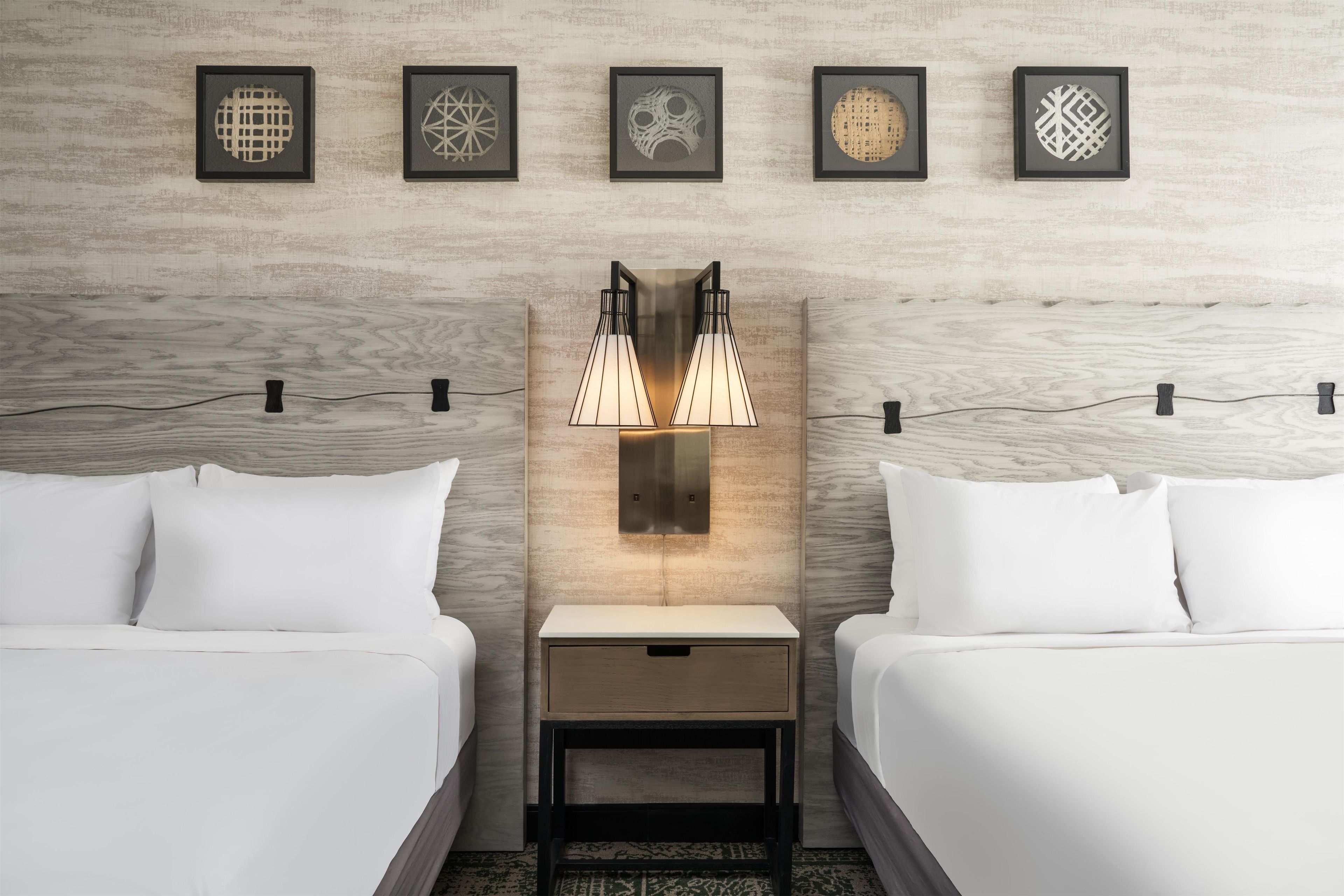 Habitación de hotel con dos camas, una mesita de noche, una lámpara de pared y cinco cuadros enmarcados.