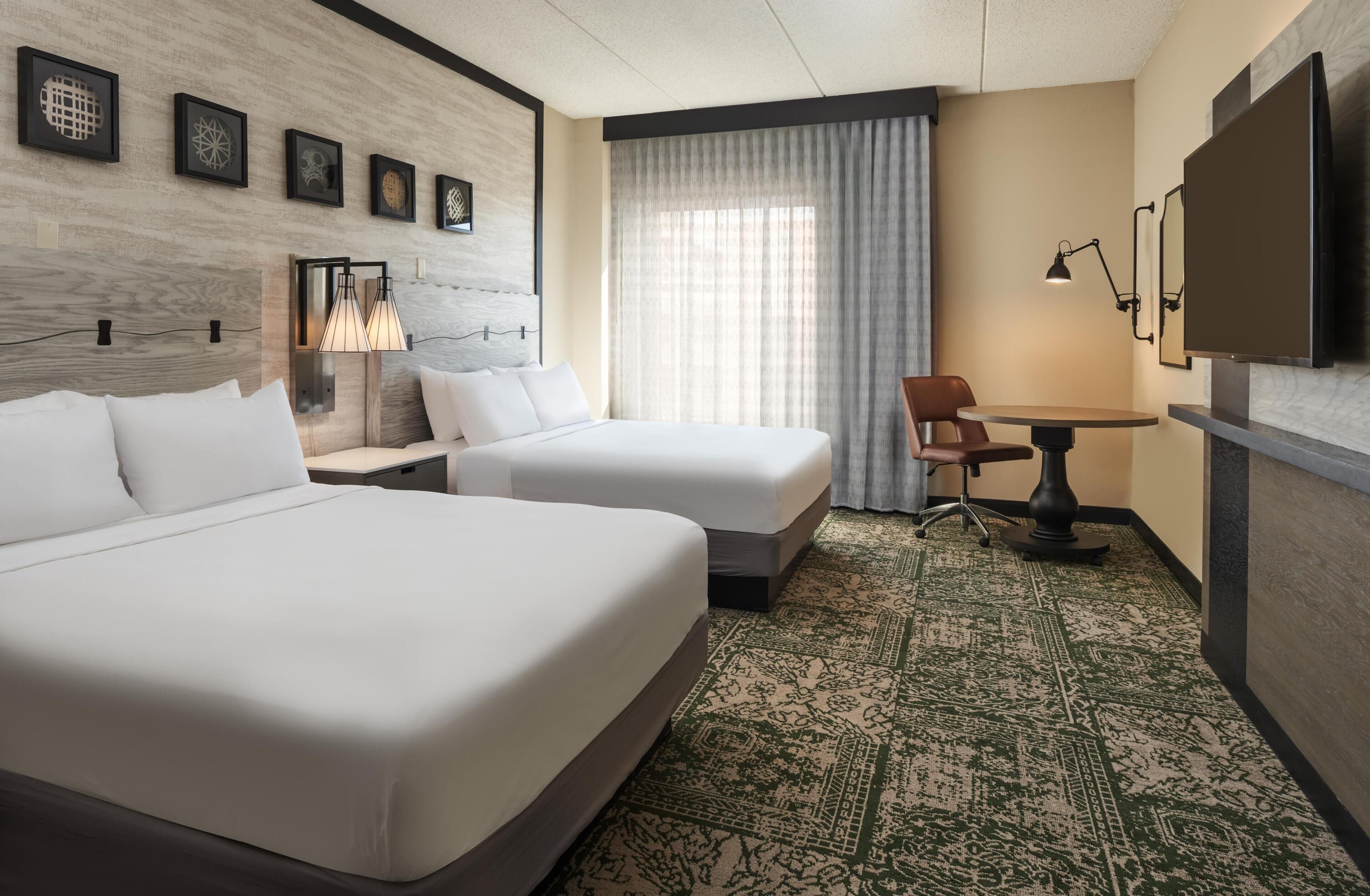 A hotel room with two beds, a desk, a chair, a TV, and a window with curtains.