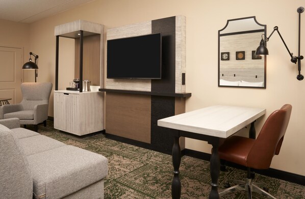A hotel room with a flat-screen TV mounted on a wall, a small table with a chair, a sofa, and a cabinet with a mirror.