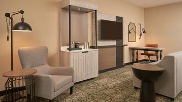 A hotel room with a grey armchair, a small round table, a cabinet with a mirror, a television mounted on the wall, and a patterned carpet.
