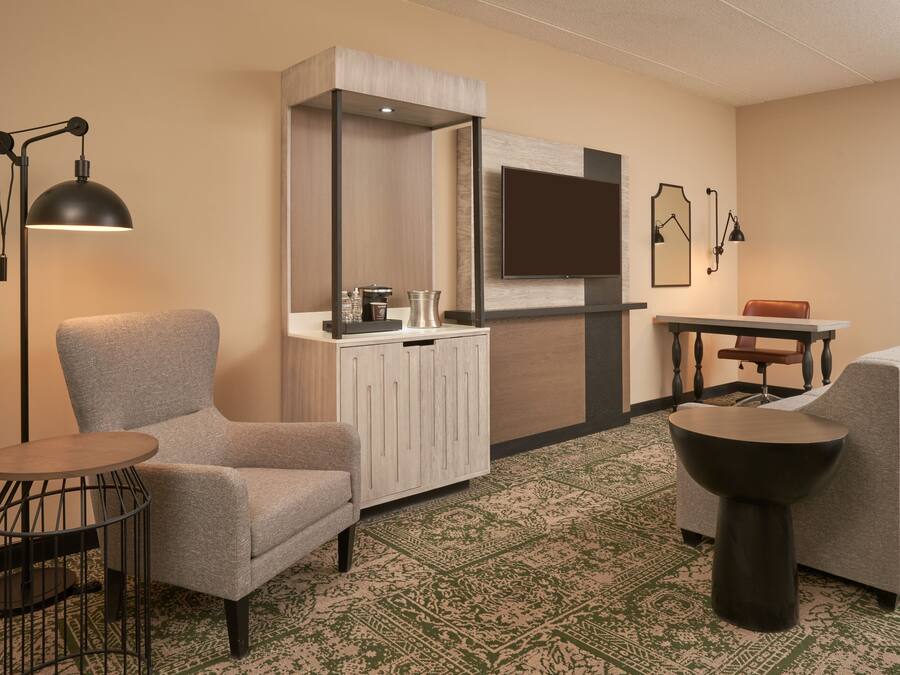 A hotel room with a grey armchair, a small round table, a cabinet with a mirror, a television mounted on the wall, and a patterned carpet.